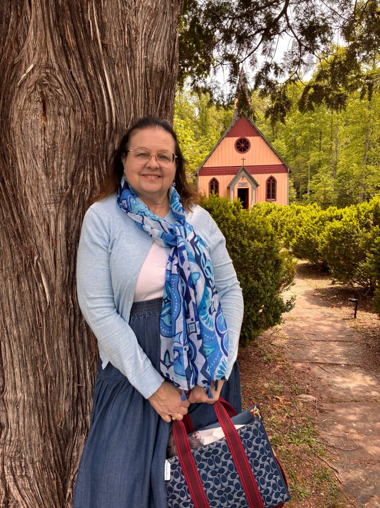 A woman stands beside a large tree, smiling while holding a handbag, with a small pink chapel and greenery in the background.