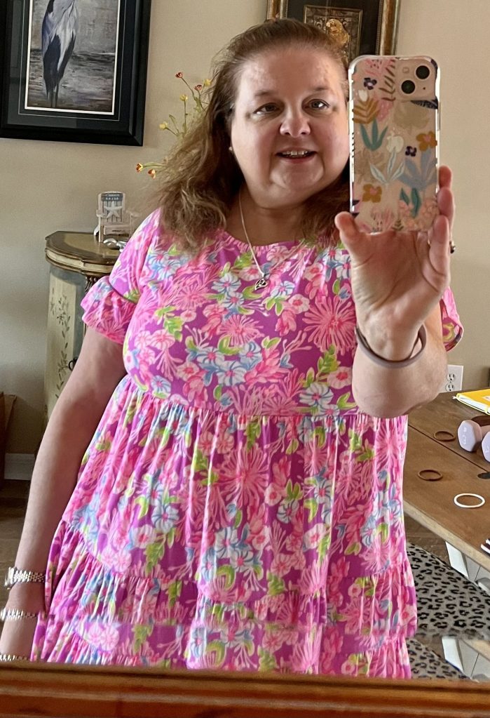 A smiling woman takes a selfie in a mirror, wearing a pink floral dress. She has long hair and is holding a phone with a patterned case. The background includes a framed picture and a small table with decorative items.