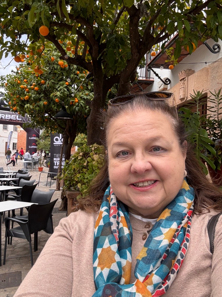 A woman smiling for a selfie in a vibrant outdoor setting, with a tree full of oranges in the background and outdoor cafe tables around her.