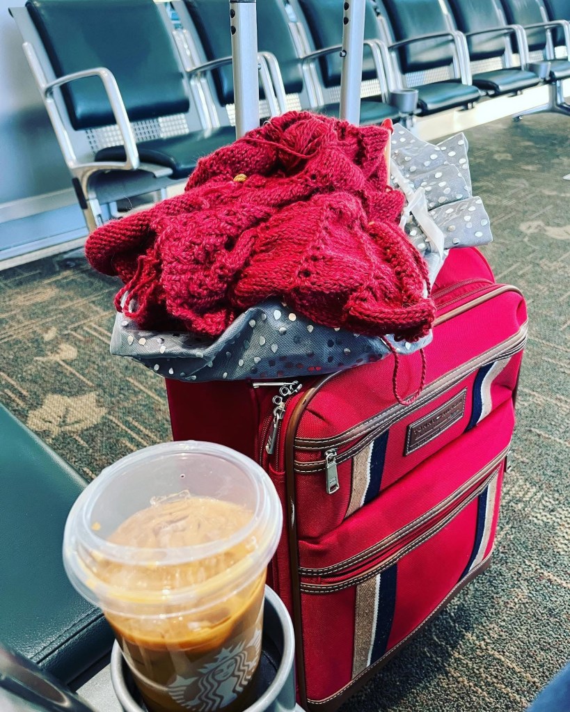 A red suitcase with a knitted sweater on top and a coffee drink in a cup holder, set in an airport waiting area.